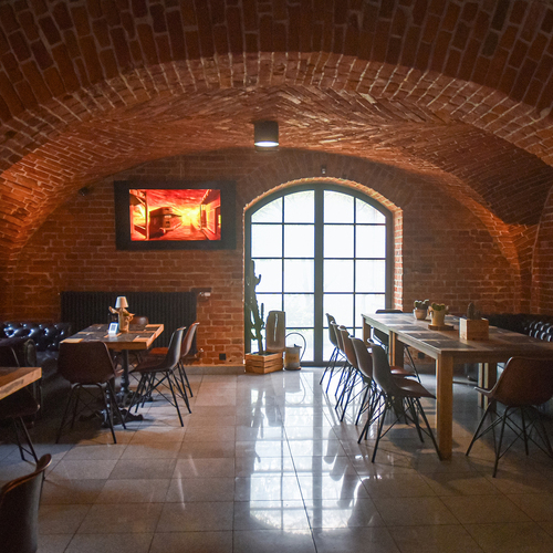 Gemütliche Lounge im gewölbten Ziegelkeller mit Bogenfenster, Ledersofas, großen Holztischen, warmer Beleuchtung und Topfpflanzen.