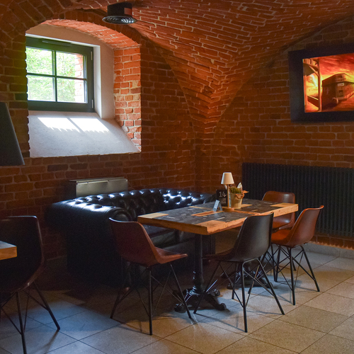 Gemütliche Hotellounge mit gewölbter Backsteindecke, Fensterlicht, Chesterfield-Ledersofa, Holztisch, Stühlen und warmer Wandkunst.