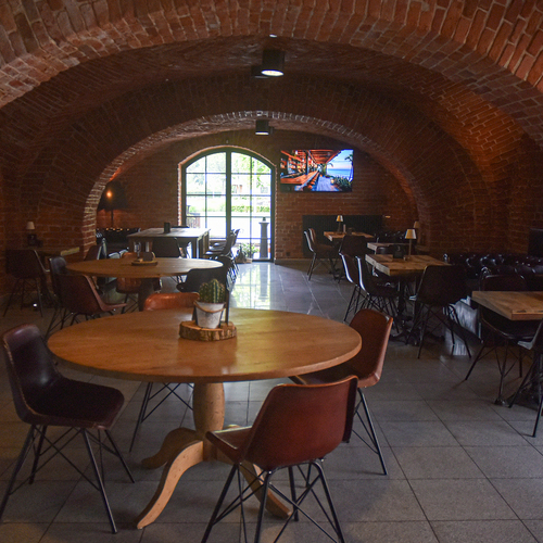 Gemütlicher Restaurantbereich in historischem Backstein-Gewölbe, runde Holztische, Lederstühle, gedämpftes Licht, Fenster und TV.
