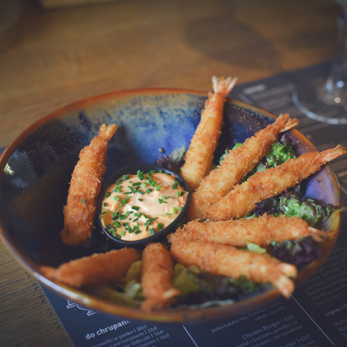 Knusprige Panko-Garnelen um eine cremige Dipsauce mit Schnittlauch in einer rustikalen blauen Keramikschale, auf einem Tisch mit Speisekarte und Weinglas.