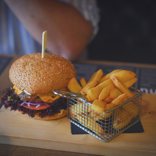 Cheeseburger mit Sesambrötchen, rotem Salat und Tomate, serviert mit goldenen Pommes im Metallkorb auf einem Holzbrett