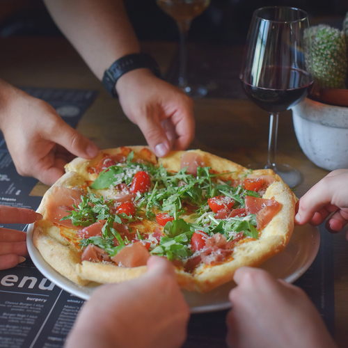 Gäste teilen eine frische Pizza mit Rucola und Prosciutto an rustikalem Tisch, Weinglas und Speisekarte sichtbar