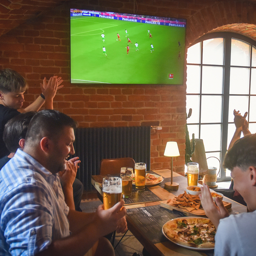 Freunde jubeln in einem gemütlichen Pub mit Backsteinwänden vor Fernseher, Pizza, Snacks und Biergläsern bei warmem Lampenlicht.