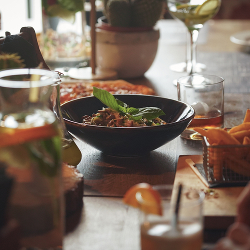Gemeinsamer Holztisch mit einer mit Basilikum garnierten Nudelschale, Pommes im Drahtkorb, Cocktails, Wein und Pizza.