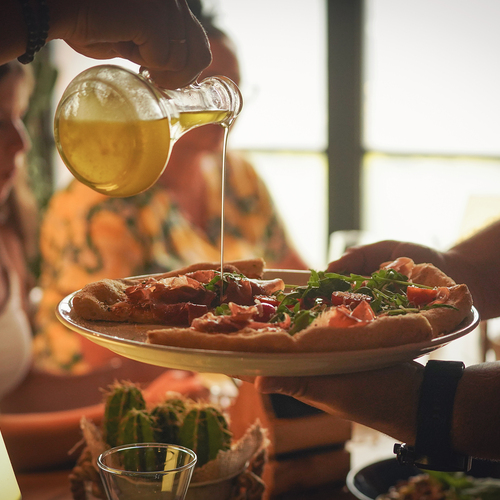 Servierender gießt goldenes Olivenöl über Prosciutto‑Rucola‑Pizza, gemütliches Zusammensitzen am Tisch