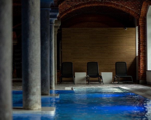 Wewnętrzny basen hotelowy z niebieską wodą, kamiennymi kolumnami, ceglanymi łukami i trzema leżakami