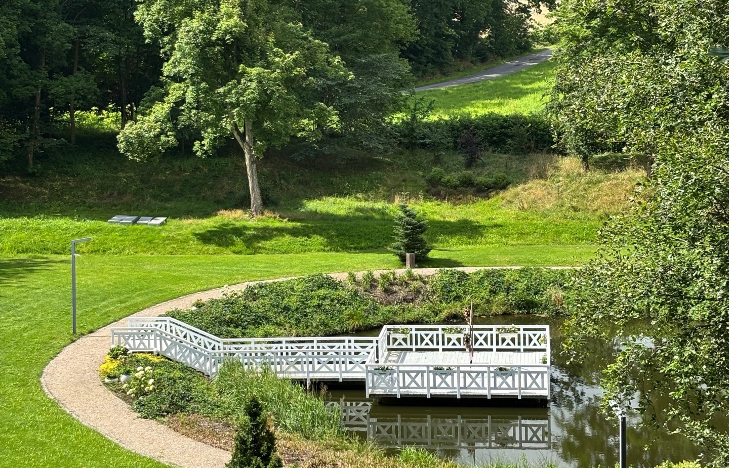 Biały drewniany pomost i pergola nad spokojnym stawem, kręte żwirowe ścieżki i zielone tereny idealne na spacery.