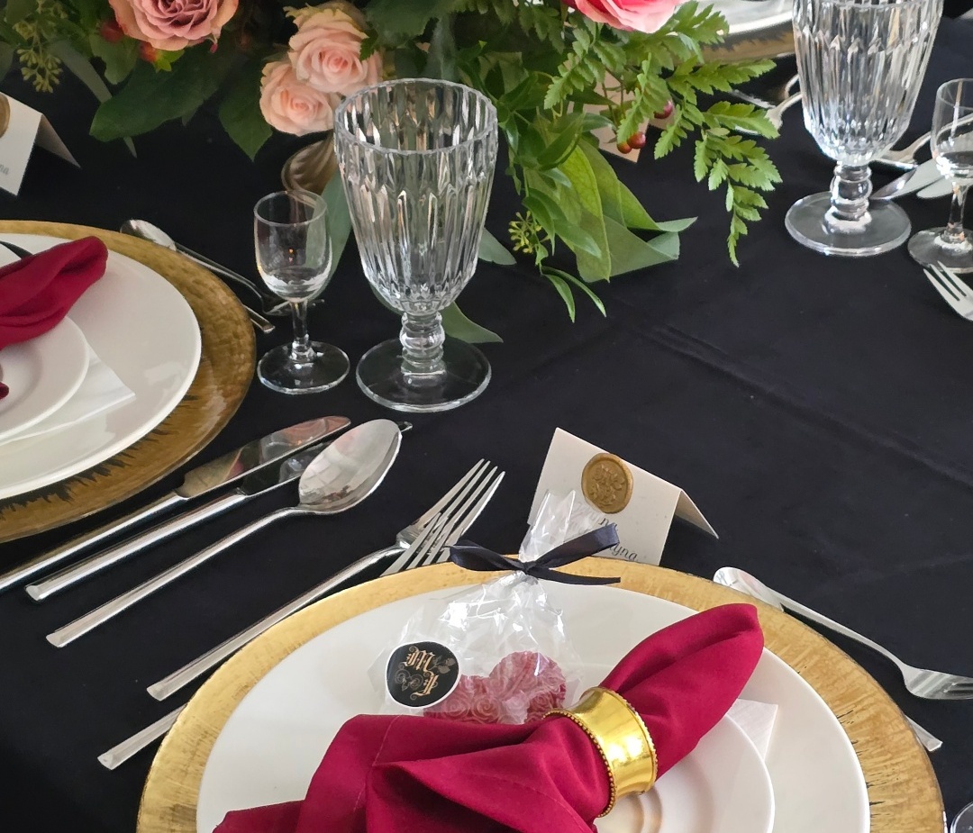 Eleganckie nakrycie stołu ze złotą podstawką, białymi talerzami, bordową serwetką w złotym pierścieniu, kryształowymi kieliszkami i bukietem róż.