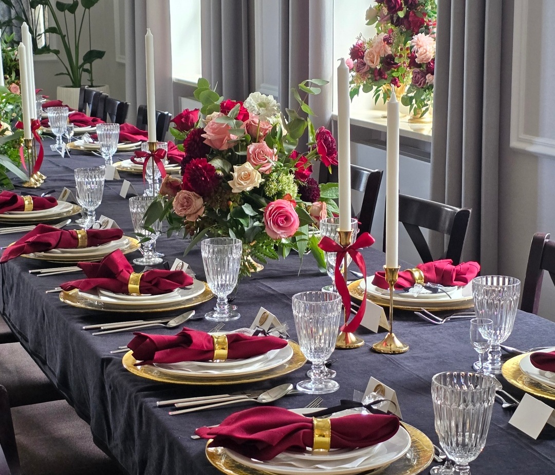 Elegancki stół bankietowy z czarnym obrusem, złotymi podtalerzami, kryształowymi kieliszkami, bordowymi serwetkami i bukietami róż przy oknach.