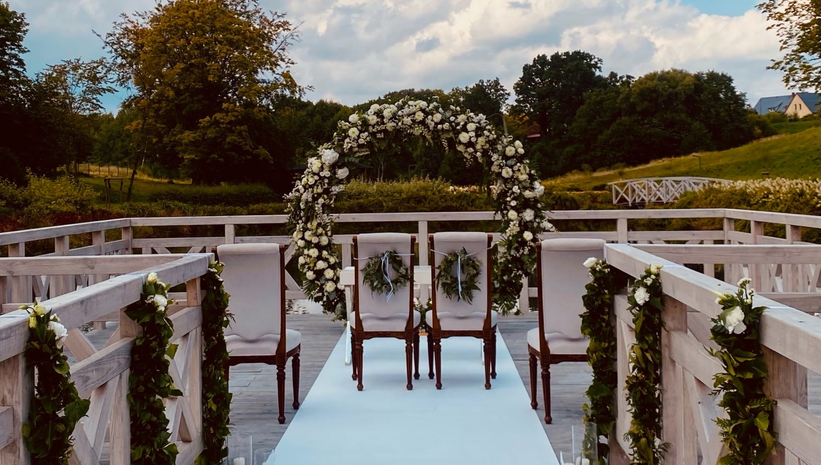 Drewniany pomost nad jeziorem na ceremonię ślubną z białym dywanem, kwiatowym łukiem, ozdobnymi krzesłami i świecami.