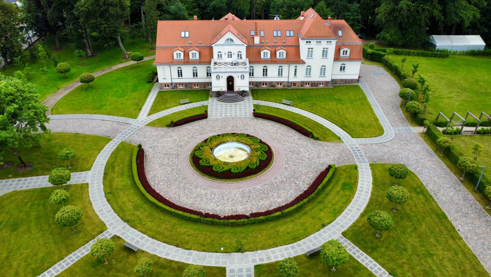 Widok z lotu ptaka na okazały dworek hotelowy z czerwoną dachówką, okrągłym brukowanym podjazdem, fontanną i zadbanymi trawnikami i alejkami.