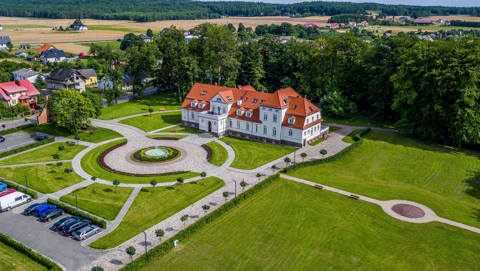 Pałacyk hotelowy w stylu manor z czerwonym dachem, brukowanym okrągłym dziedzińcem z fontanną, zadbanymi trawnikami, parkingiem i zalesieniem w tle