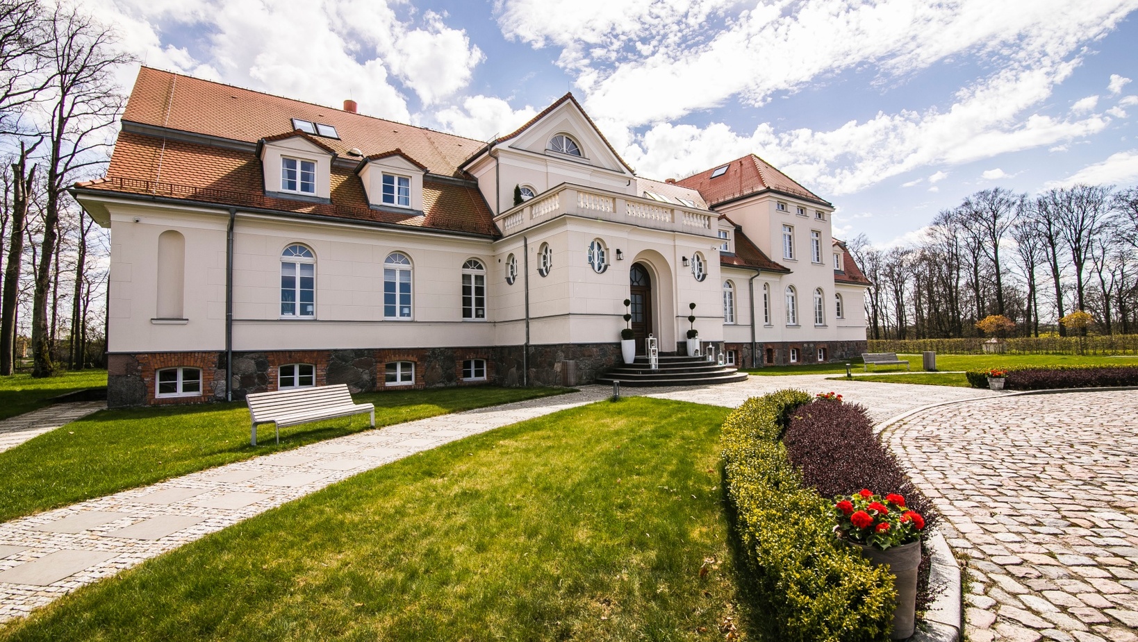 Elegancki hotel-pałac na wsi, kremowa elewacja, czerwony dach, łukowe wejście, brukowany podjazd, zadbany trawnik i donice z czerwonymi kwiatami.