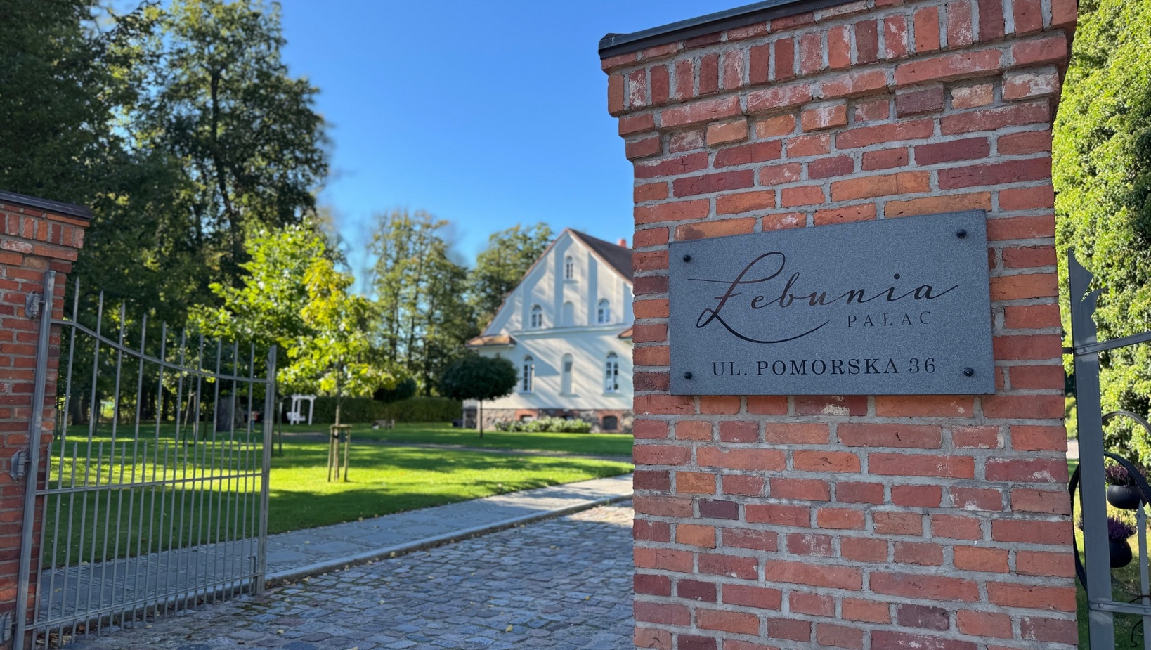 Biały pałacyk za ceglanym wejściem z otwartą metalową bramą, kostkowana aleja i zadbany trawnik