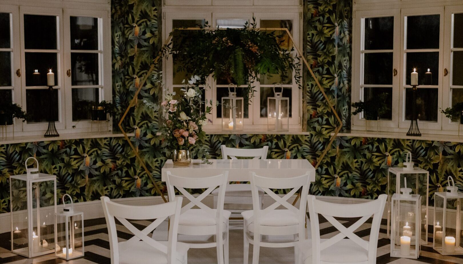 Kameralny stół ceremonii z białymi krzesłami, kwiatową dekoracją pod złotą geometryczną arkadą, lampiony z świecami i tropikalna tapeta