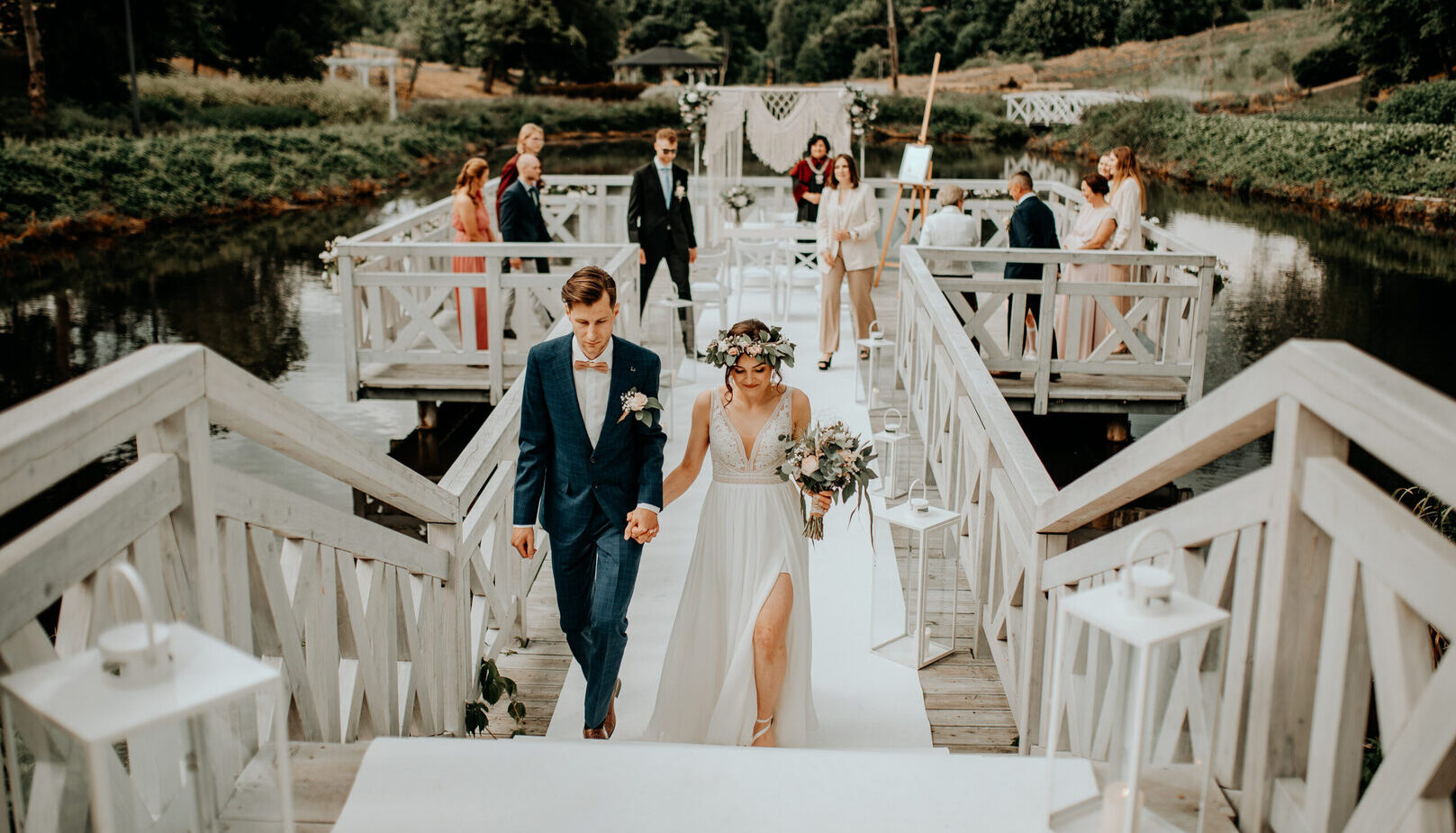 Para młoda idzie za rękę po białym drewnianym pomoście nad jeziorem, z makramowym łukiem, gośćmi i latarniami