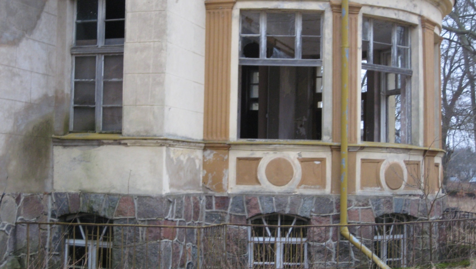 Zniszczona kamienna i tynkowa elewacja dawnej willi z wybitymi oknami, półokrągłym wykuszem i żółtą rynną