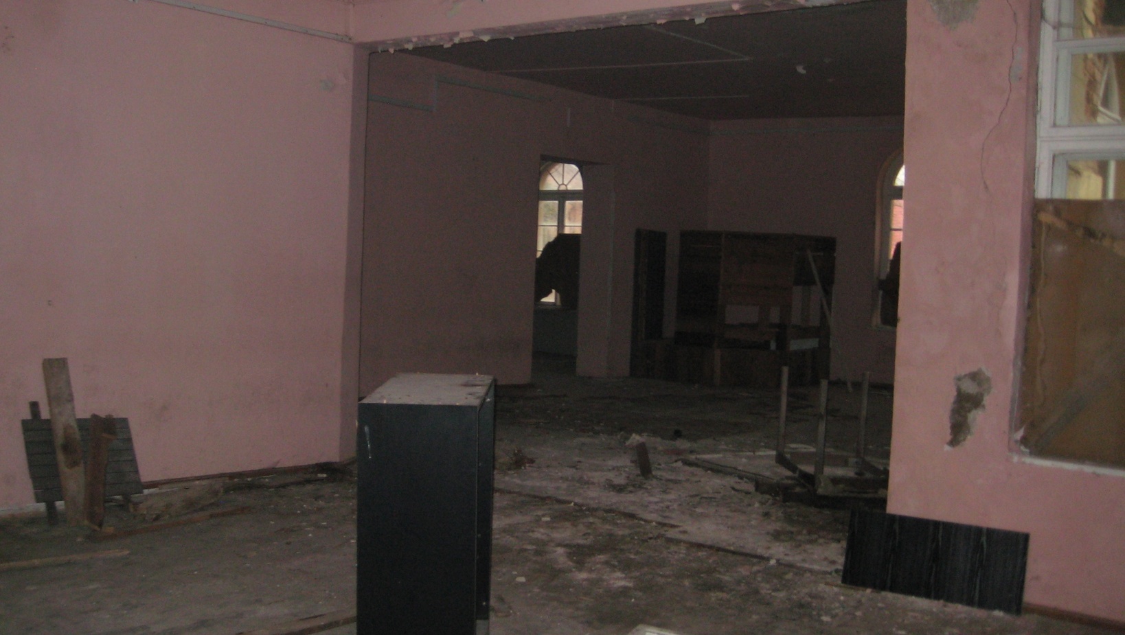 Opuszczone, zakurzone wnętrze z wyblakłymi różowymi ścianami, porozrzuconym gruzem, zniszczonymi meblami i łukowymi oknami.