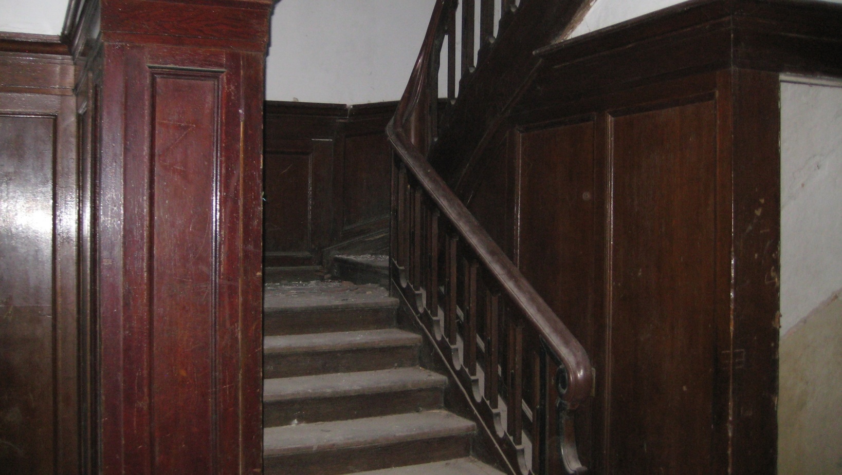 Zniszczone drewniane schody z rzeźbioną balustradą i ciemnymi boazeriami w zabytkowym, przytłumionym korytarzu