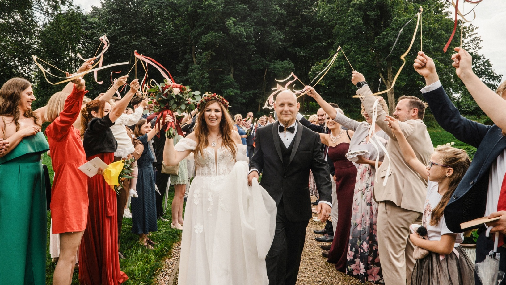 Panna młoda w koronkowej białej sukni z wiankiem i pan młody w czarnym smokingu idą przez alejkę gości machających wstążkami.