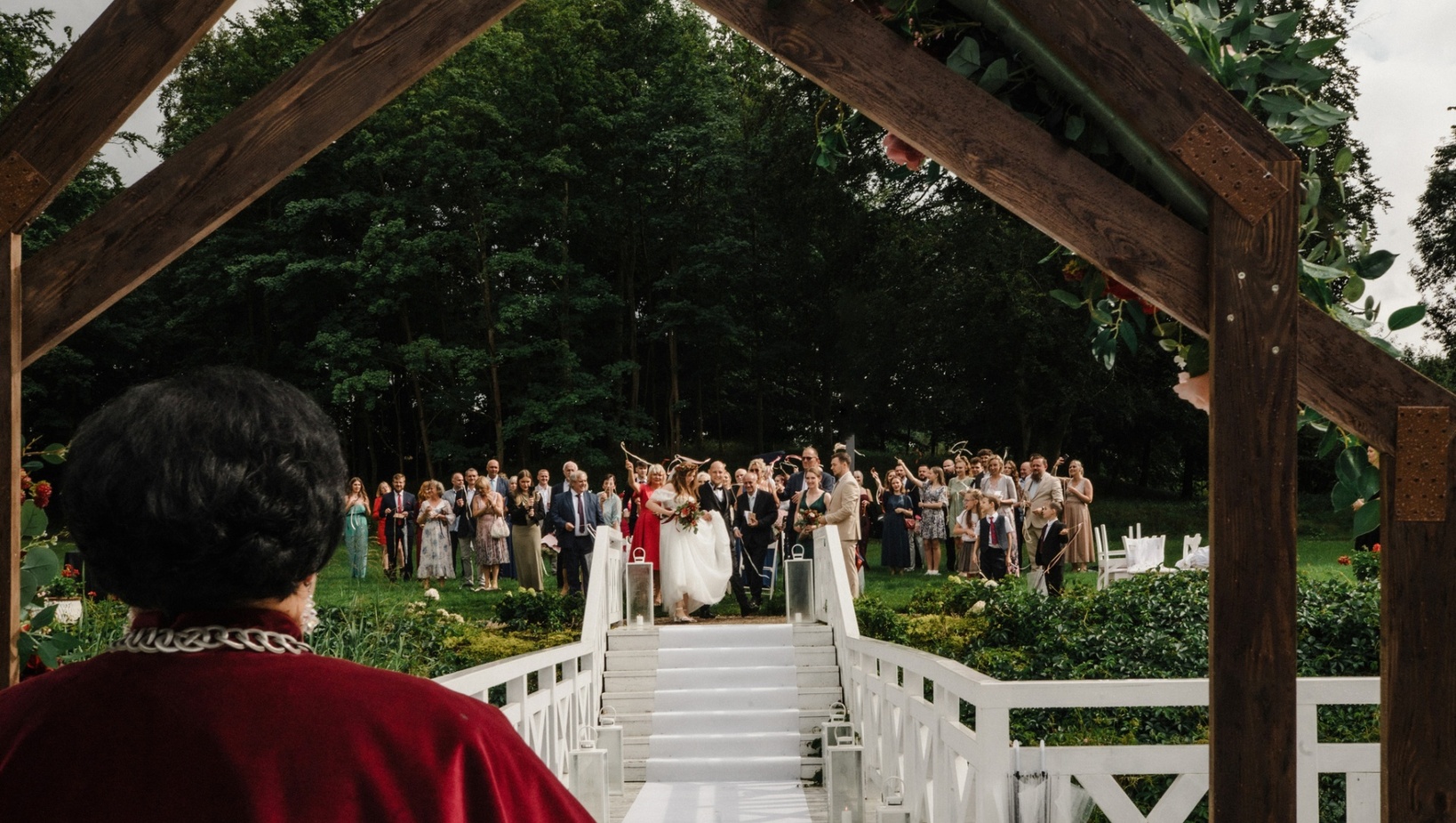 Plenerowy ślub na białym moście z drewnianą arkadą ozdobioną kwiatami, para młoda schodzi alejką, goście wiwatują.
