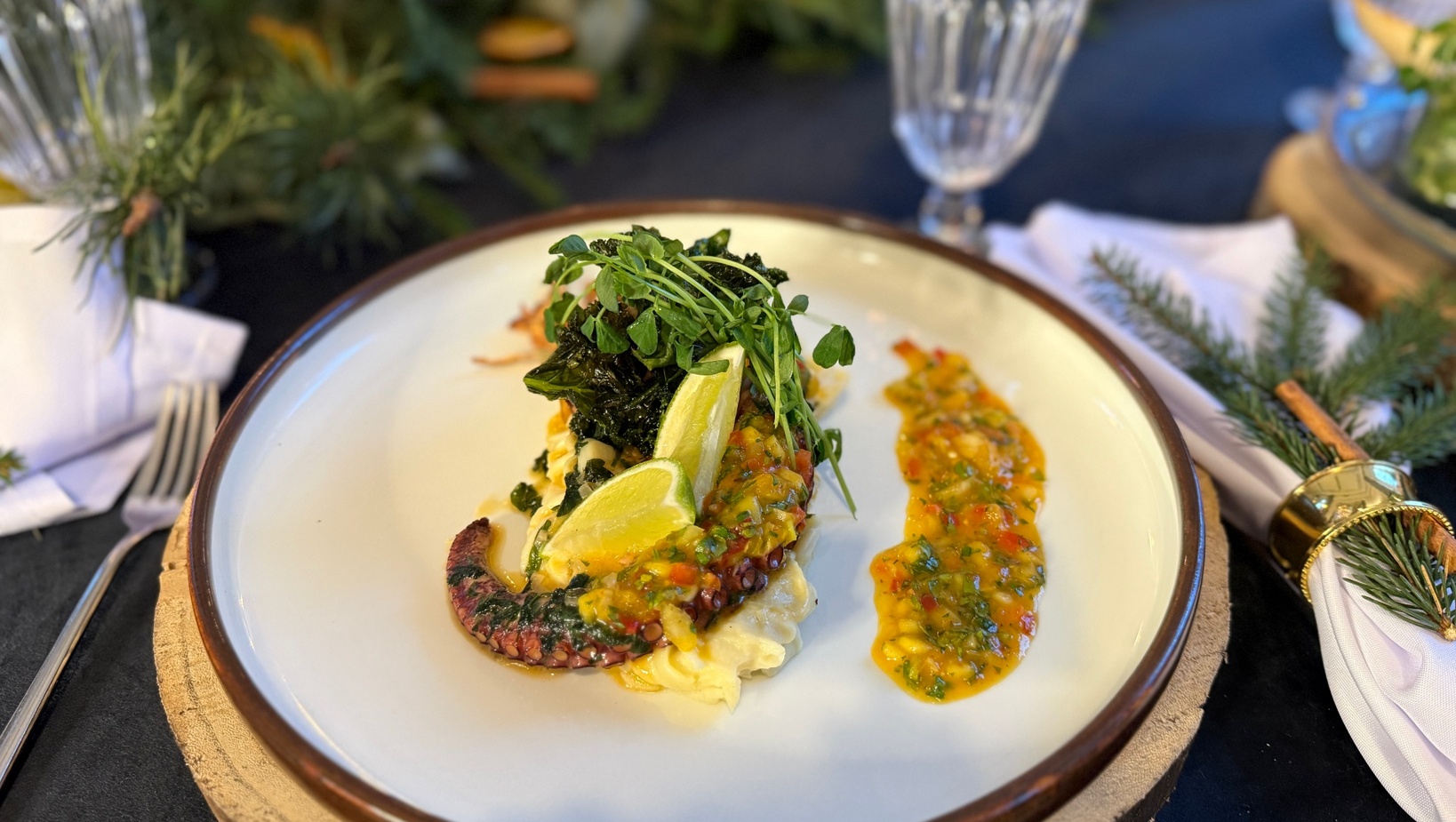 Grillowana ośmiornica na kremowym purée z ćwiartkami cytrusów, salsą z ziół i kiełkami, podana na rustykalnym talerzu obok kryształowych kielichów.