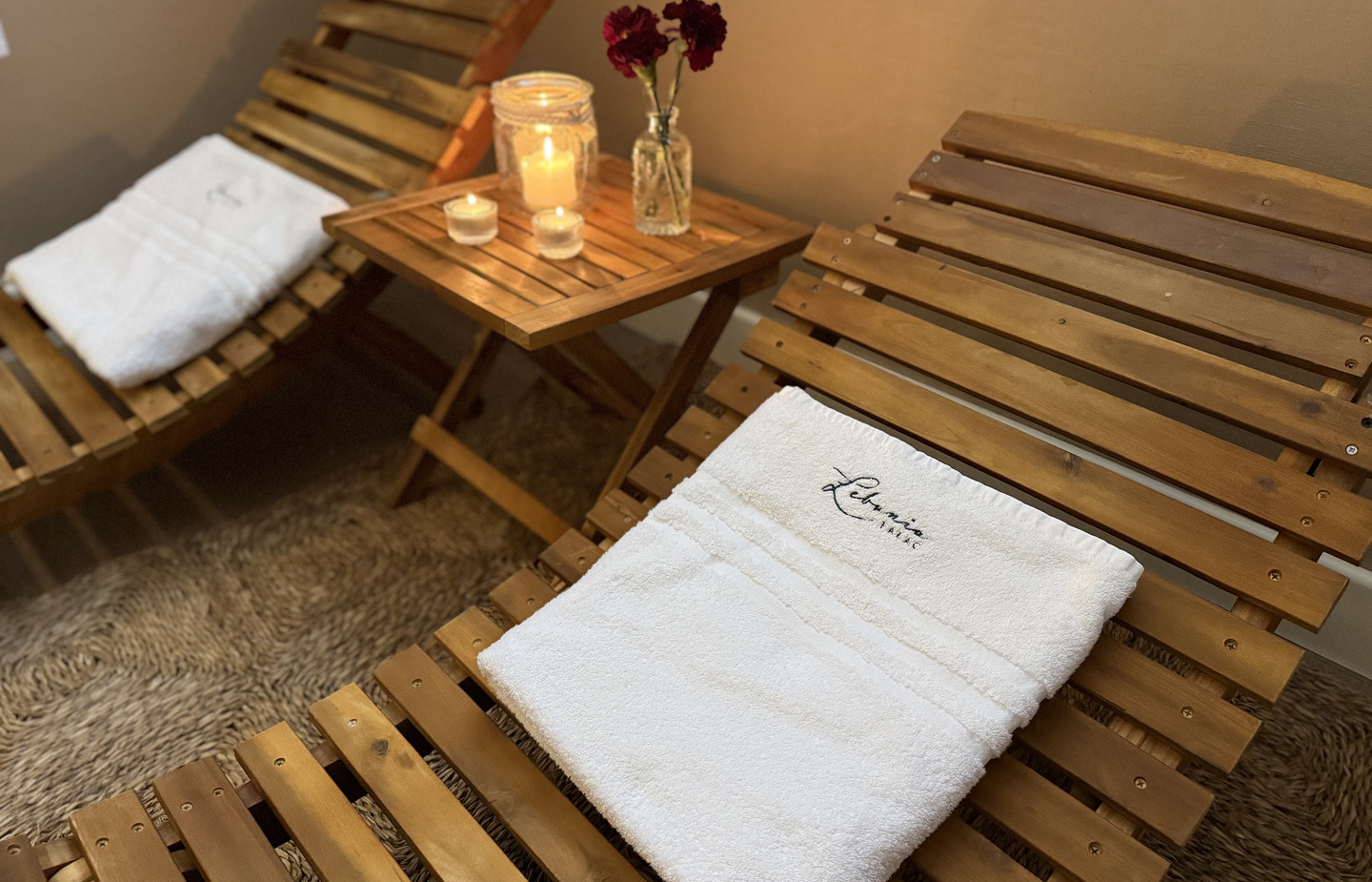 Drewniane leżanki z białymi hotelowymi ręcznikami, stolik z zapalonymi świecami i czerwonymi kwiatami, przytulna atmosfera.