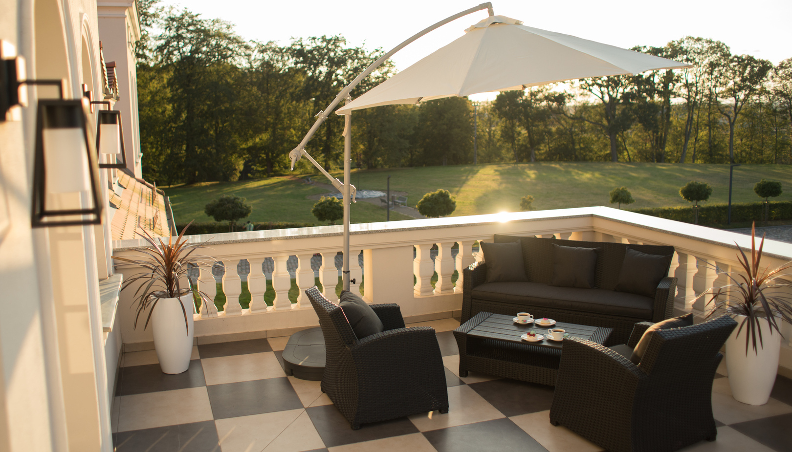 Taras hotelowy w słońcu z rattanową sofą i fotelami, stolikiem z kawą, parasolem i widokiem na trawnik.