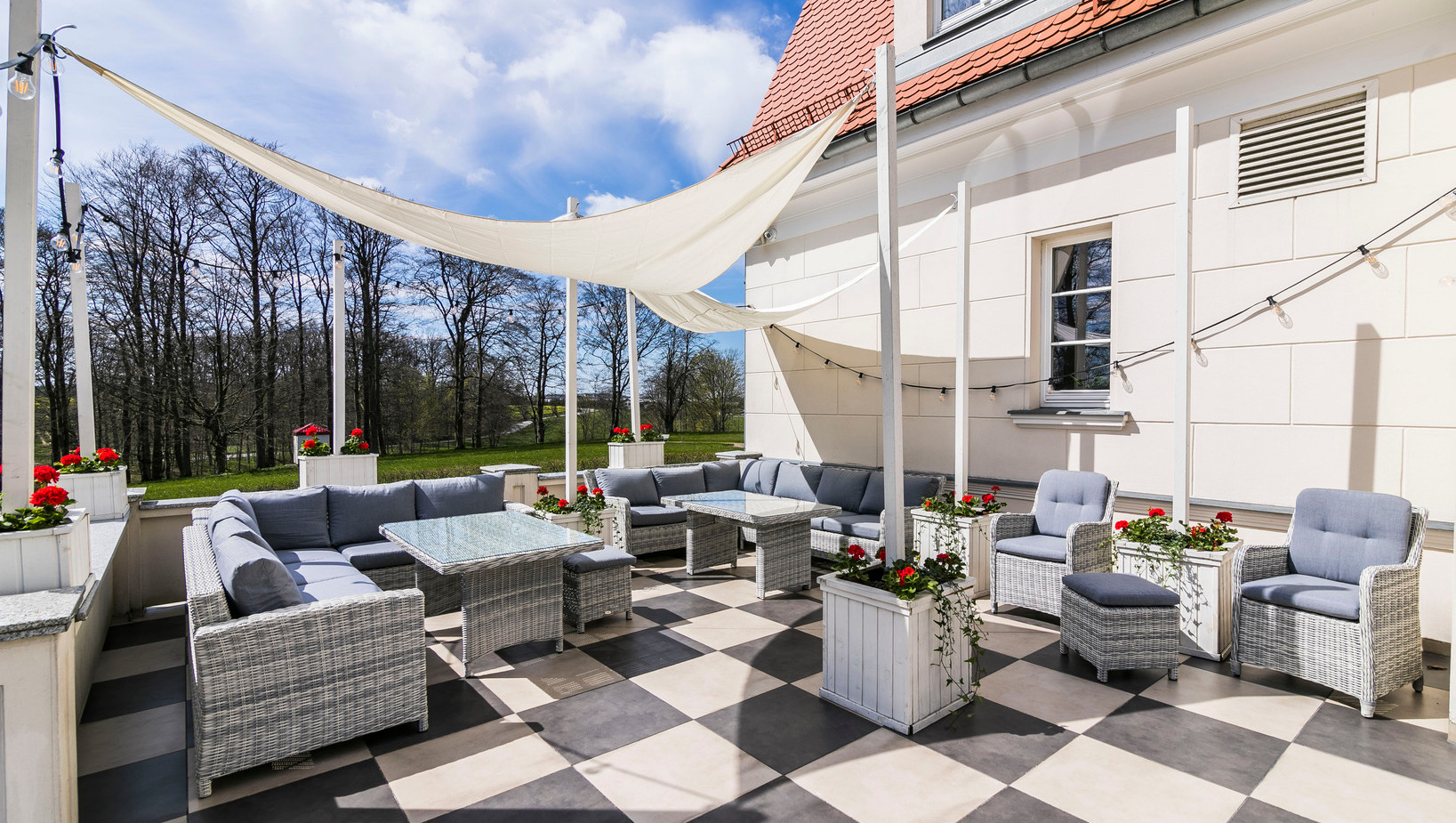 Słoneczny taras hotelowy z plecionymi sofami i fotelami, szklanymi stolikami, żaglami przeciwsłonecznymi, czarno-białą posadzką i pelargoniami.