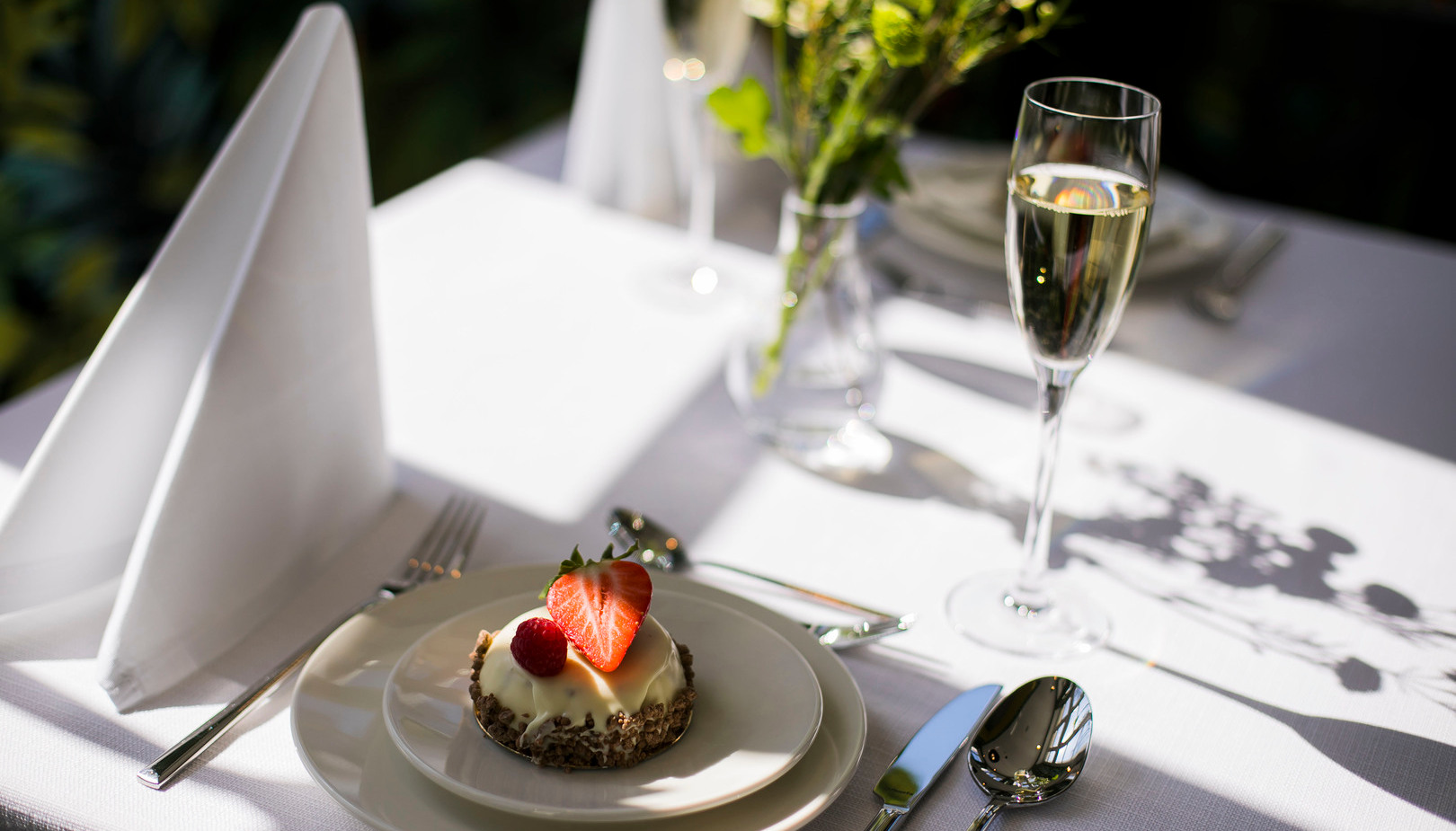 Stolik z białym obrusem w restauracji: deser z truskawką i maliną na talerzach, kieliszek szampana, złożona serwetka i wazon z kwiatami.