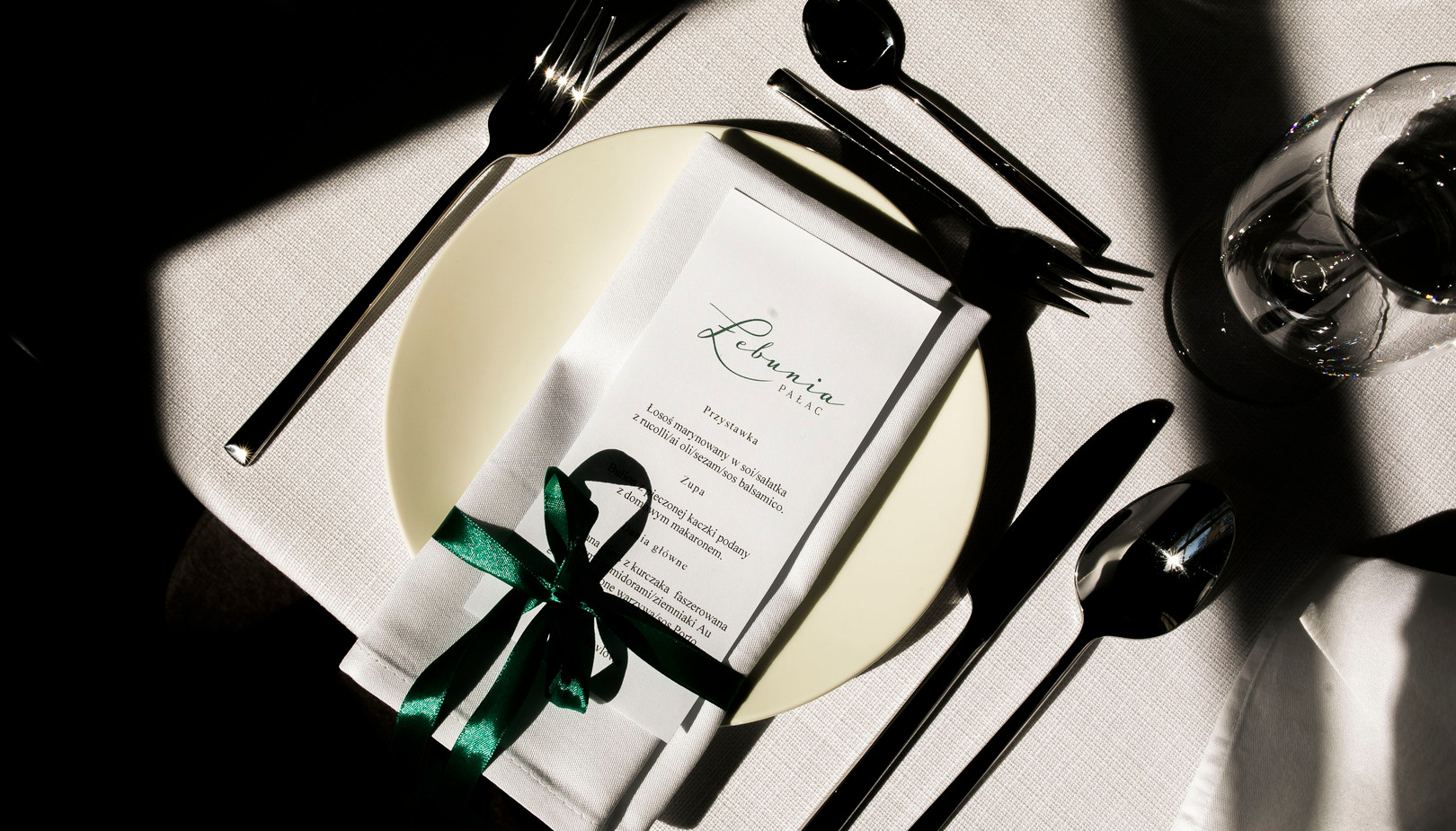 Elegancka aranżacja stołu: biały talerz z serwetką, menu związane zieloną wstążką, błyszczące sztućce i kieliszek.
