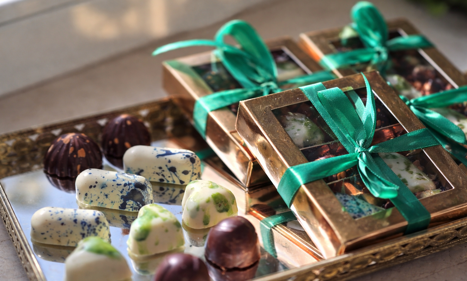 Porcja ekskluzywnych pralinek na lustrzanej tacy, białe i zielone kropkowane czekoladki obok złotych pudełek przewiązanych zieloną wstążką