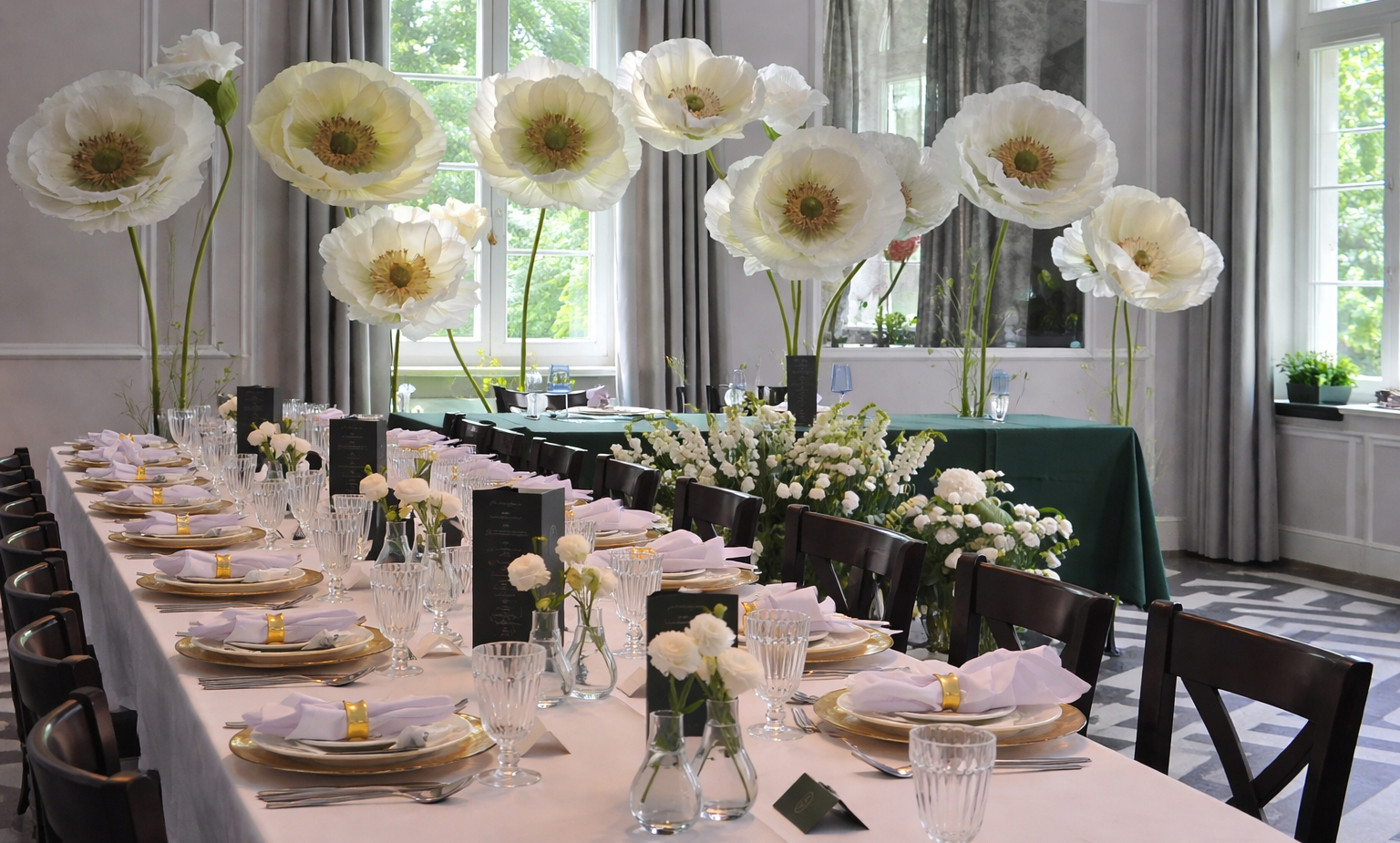 Bankietowy stół z złotymi podkładkami, kryształowymi kieliszkami, białymi serwetkami i dużymi białymi kwiatami dekoracyjnymi.