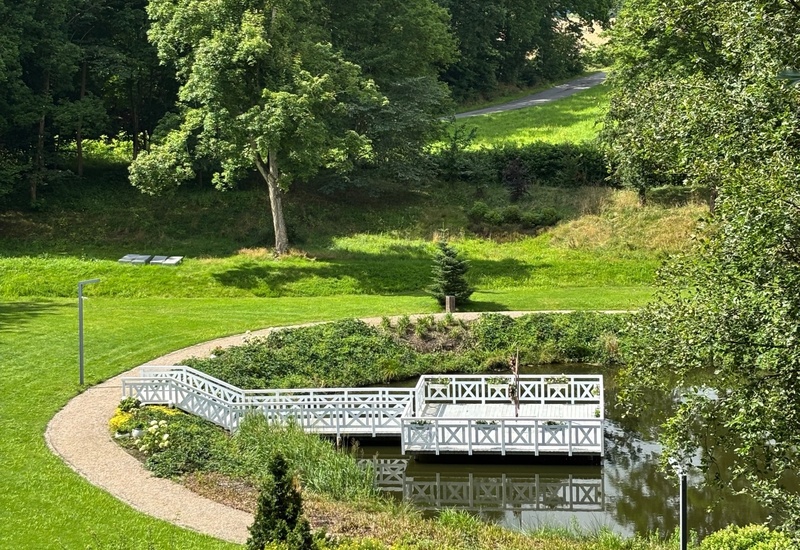 Biały drewniany pomost i pergola nad spokojnym stawem, kręte żwirowe ścieżki i zielone tereny idealne na spacery.