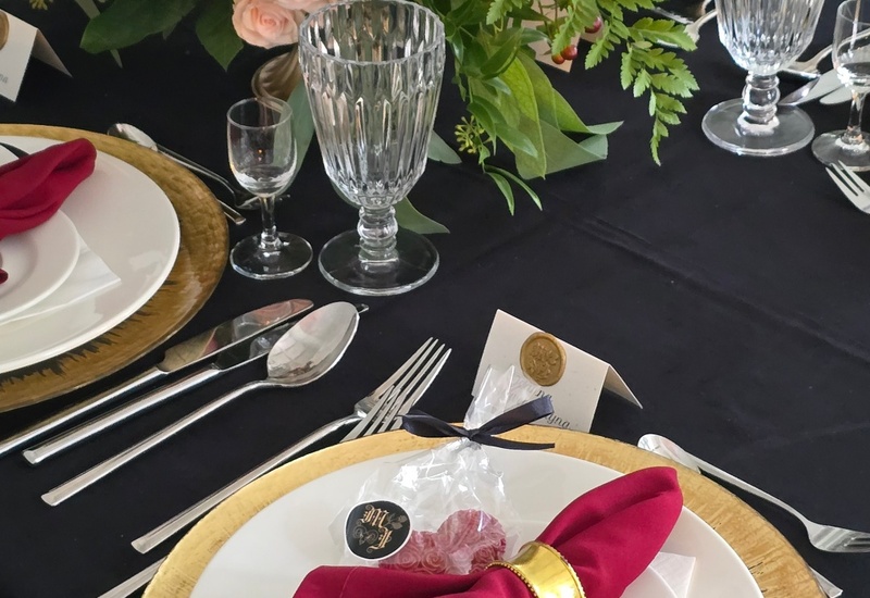 Eleganckie nakrycie stołu ze złotą podstawką, białymi talerzami, bordową serwetką w złotym pierścieniu, kryształowymi kieliszkami i bukietem róż.