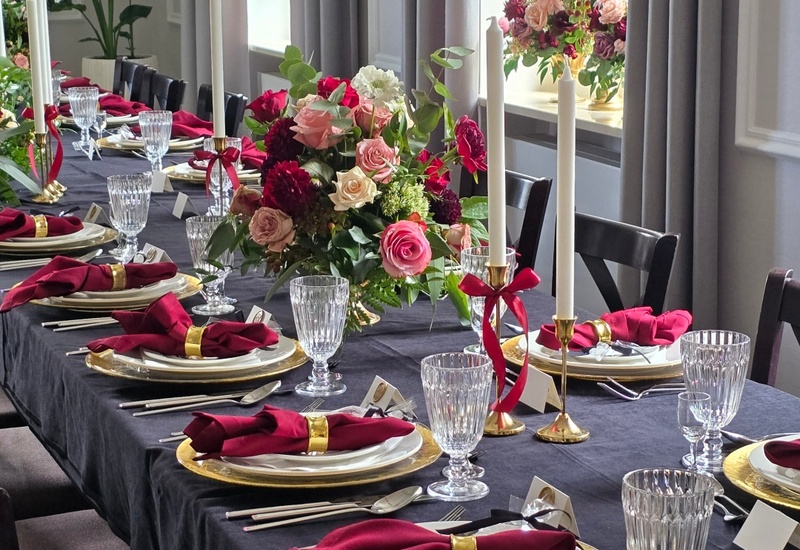 Elegancki stół bankietowy z czarnym obrusem, złotymi podtalerzami, kryształowymi kieliszkami, bordowymi serwetkami i bukietami róż przy oknach.
