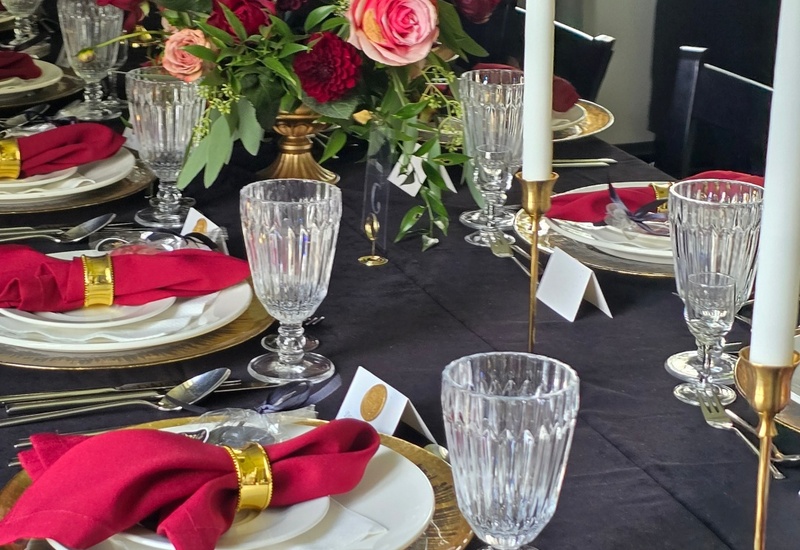 Elegancki stół z czarnym obrusem, złotymi podtalerzami, czerwonymi serwetkami w złotych obrączkach, kryształowe szkło, róże