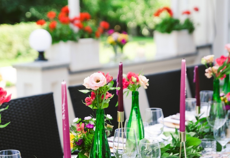 Stolik na tarasie przygotowany na posiłek: kwiatowe bukiety, zielone butelki, fioletowe świece i szkło
