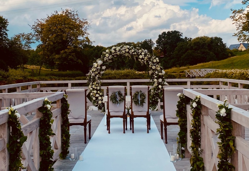 Drewniany pomost nad jeziorem na ceremonię ślubną z białym dywanem, kwiatowym łukiem, ozdobnymi krzesłami i świecami.