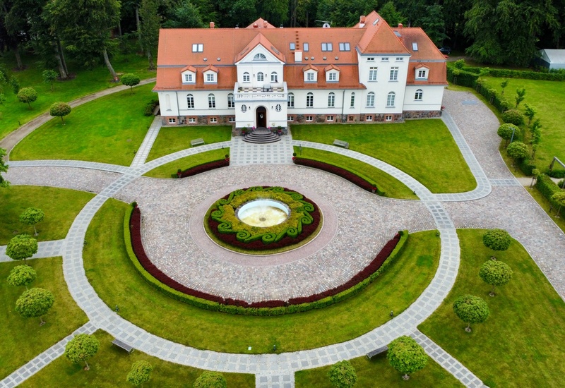 Widok z lotu ptaka na okazały dworek hotelowy z czerwoną dachówką, okrągłym brukowanym podjazdem, fontanną i zadbanymi trawnikami i alejkami.