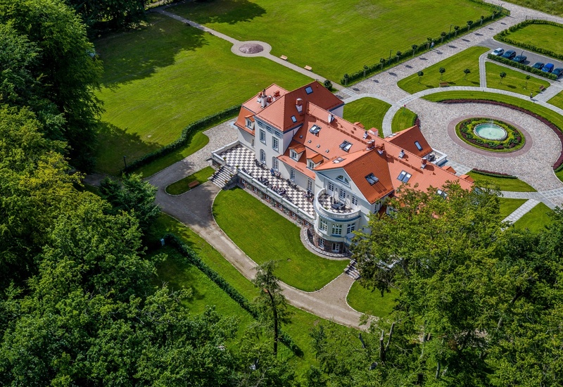 Hotel butikowy z czerwoną dachówką, tarasem w szachownicę, okrągłym podjazdem z fontanną oraz zadbanymi trawnikami i parkiem.