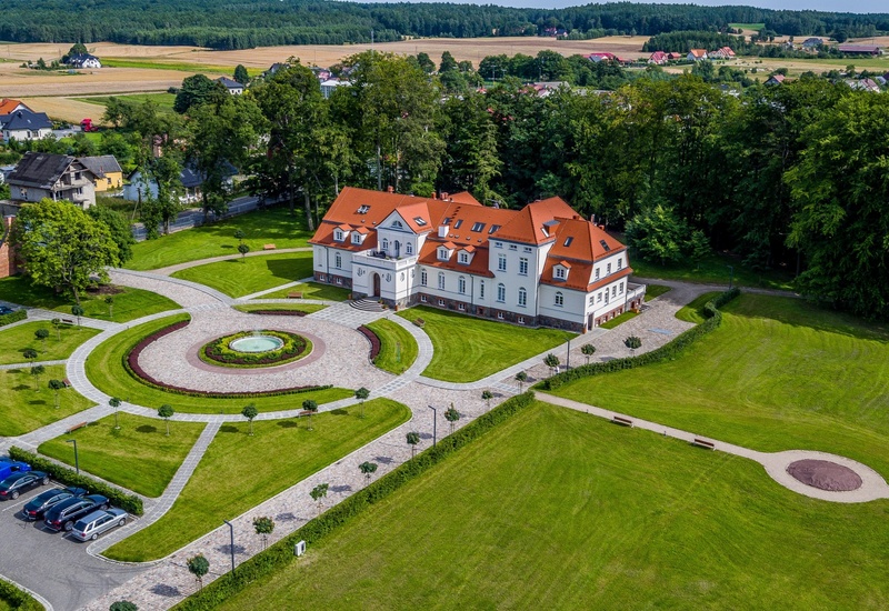 Pałacyk hotelowy w stylu manor z czerwonym dachem, brukowanym okrągłym dziedzińcem z fontanną, zadbanymi trawnikami, parkingiem i zalesieniem w tle