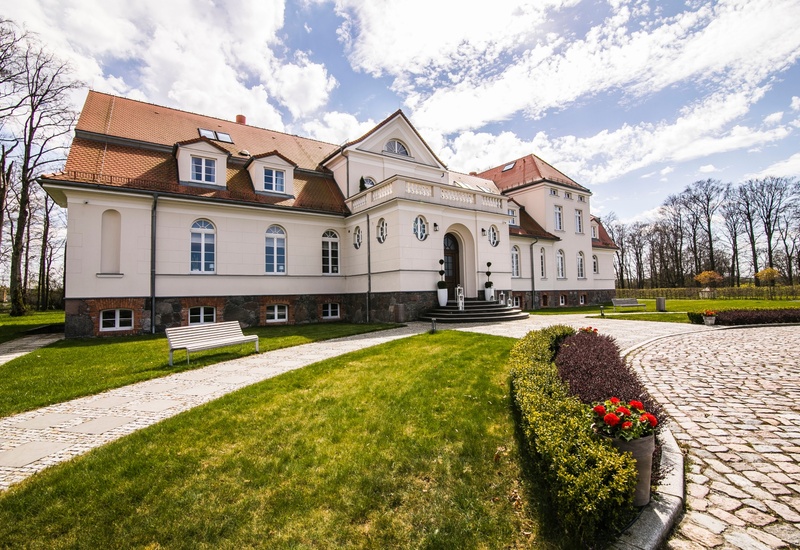 Elegancki hotel-pałac na wsi, kremowa elewacja, czerwony dach, łukowe wejście, brukowany podjazd, zadbany trawnik i donice z czerwonymi kwiatami.