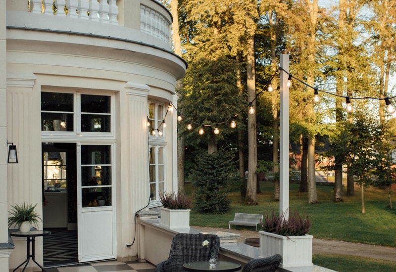 Elegancki taras hotelu z szachownicową podłogą, fotelami i stolikiem, lampkami i widokiem na park o zachodzie