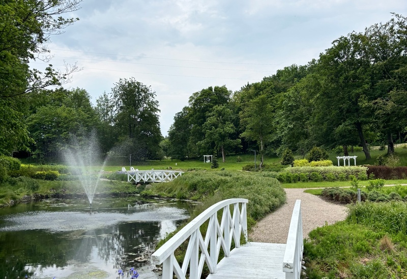Biały drewniany mostek nad stawem z fontanną, żwirowe ścieżki i fioletowe irysy w zadbanym parku idealnym na spacer.