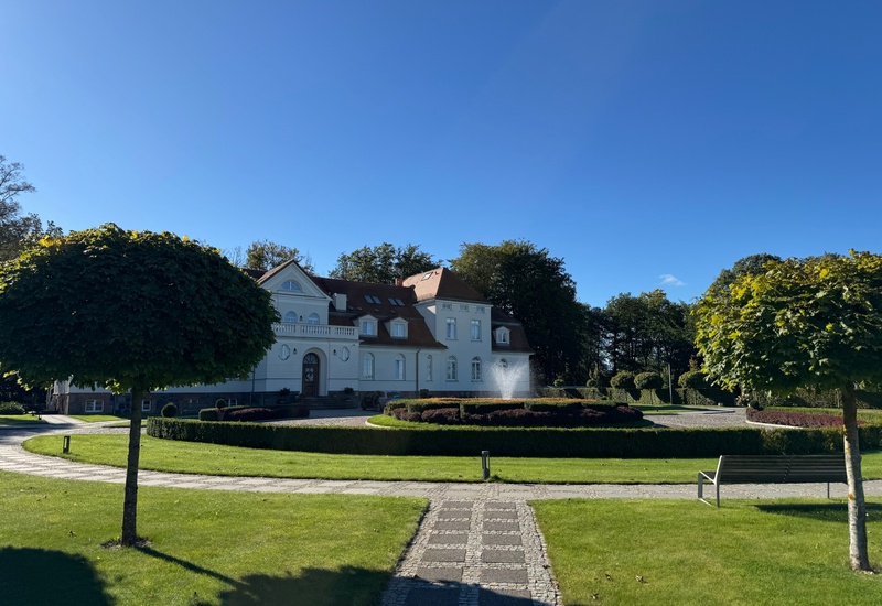 Historyczny biały dwór z funkcją hotelu, zadbane trawniki, centralna fontanna, przycięte drzewka, brukowana ścieżka i ławki