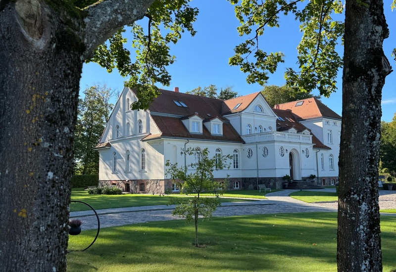 Biały pałacyk z czerwonym dachem, reprezentacyjnym wejściem, brukowanym podjazdem i zadbaną trawą wśród dojrzałych drzew