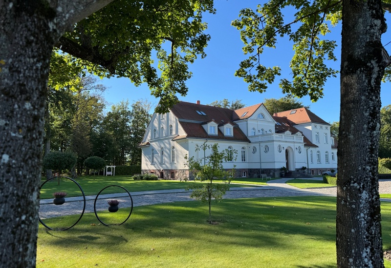 Biała rezydencja hotelowa z czerwoną dachówką, balkonem i brukowanym podjazdem, otoczona drzewami i zadbanym trawnikiem.