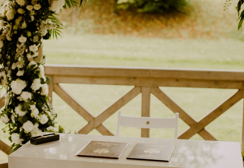Biały stół do podpisów z dwoma dokumentami pod bujną kwiatową arkadą w drewnianej altanie, plener ślubny