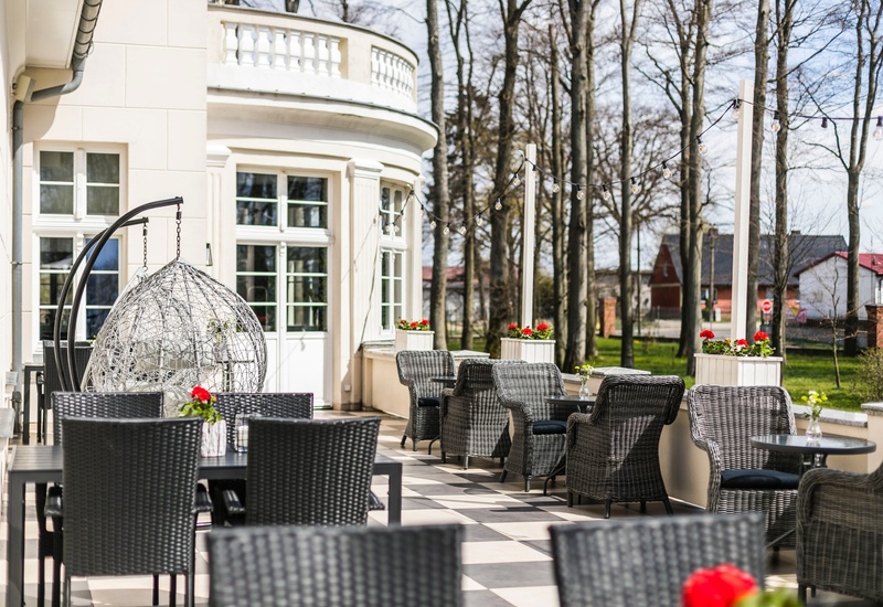 Słoneczny taras hotelowy z wiklinowymi fotelami, wiszącym kokonem, girlandą świateł i donicami z czerwonymi pelargoniami